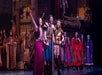 Performers in medieval-style costumes take a bow on stage, with a detailed backdrop and other cast members in the background.