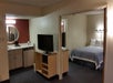 A hotel room suite with a bed in a separate sleeping area, a TV on a stand, and a small kitchenette with a mirror and sink.