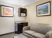 Hotel room with a plaid couch, kitchen area including a microwave and mini-fridge, and landscape artwork on cream-colored walls. A floor lamp stands beside the couch.