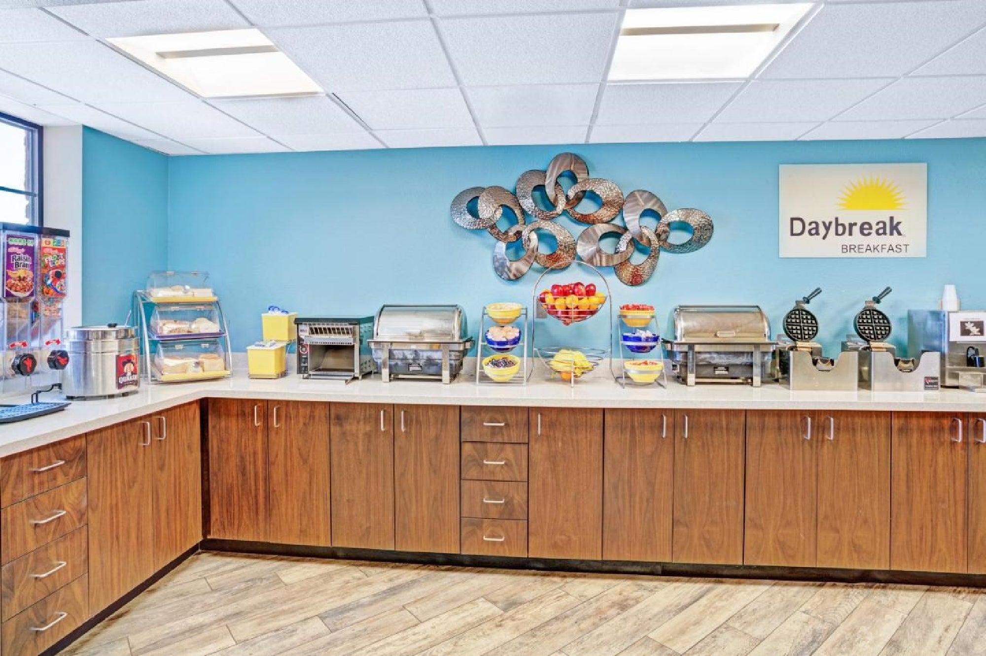 Hotel breakfast buffet area with a counter featuring cereal dispensers, fruit bowls, waffle makers, and various food warmers. A "Daybreak Breakfast" sign is on the blue wall above.