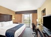 Hotel room with a large bed, dark headboard, desk, chair, TV, and large window with curtains. Light-colored walls and carpeted floor.