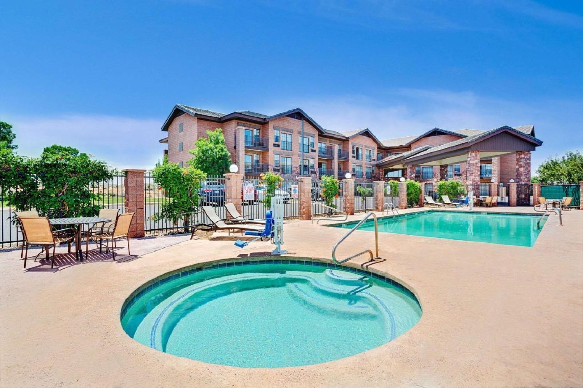 Outdoor pool area with a round hot tub in the foreground and a rectangular swimming pool in the background. Apartments and patio furniture are visible under a clear blue sky.