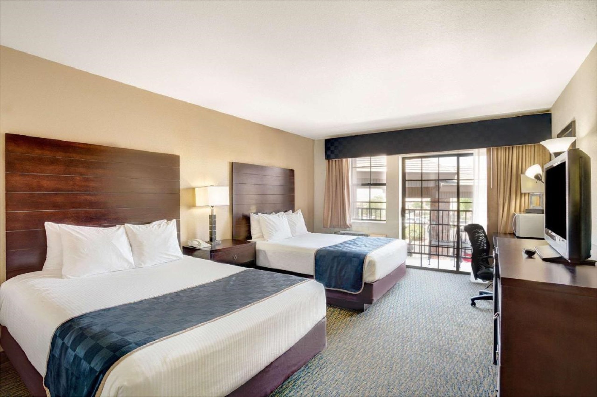Hotel room with two double beds, white linens, and dark wood headboards. A desk, chair, TV, and lamp are present. The room has large windows with curtains and a view of a balcony.