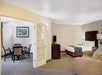 Hotel suite interior with a bedroom featuring a bed, TV, and armchair. Adjacent room has a dining table and chairs. Walls are light-colored with a framed picture and lamps providing lighting.