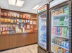 A small convenience store corner with shelves of snacks and a refrigerated section stocked with drinks like water, soda, and energy drinks.
