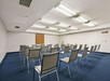 A small conference room with rows of empty gray chairs facing a blank corkboard on a white wall, under fluorescent lighting on a blue carpeted floor.