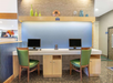 Two green chairs at a wooden desk with two computer monitors in a hotel lobby area, decorated with colorful vases on a shelf above.