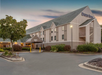A three-story hotel building with a sign that reads "Days Inn." The entrance is surrounded by landscaping, and a large parking area is in the foreground. The sky is a soft evening gradient.