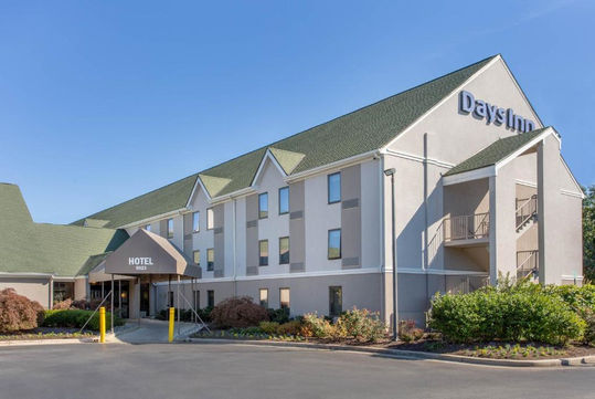 Three-story hotel building with a green roof, beige exterior, and a "Days Inn" sign. The entrance has a canopy labeled "HOTEL," surrounded by landscaped bushes and a driveway.