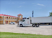 A white semi-truck is parked in front of a two-story motel with red roofs and a white sedan nearby. The sky is clear and blue.