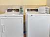 A commercial washer and dryer are side by side in a laundry room. The washer has an open detergent lid, and the dryer has a handle visible on the front.