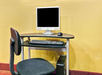 A small computer desk with a monitor, keyboard, and mouse. A black chair is positioned in front of the desk. The background is a plain yellow wall.
