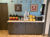 Coffee station with three large coffee dispensers, stacks of cups, creamers, sugar packets, a microwave, and a napkin dispenser on a countertop. Two framed signs are on the wall above.