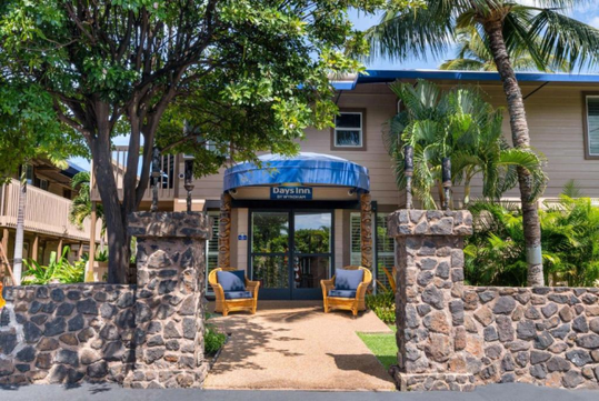 Entrance to a Days Inn hotel with stone pillars, greenery, palm trees, and two wicker chairs by the glass doors under a blue awning.