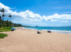 Sandy beach with a few umbrellas and chairs, bordered by green vegetation and palm trees, under a blue sky with scattered clouds near a calm ocean.