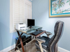 A small home office setup with a glass desk, computer monitor, keyboard, printer, and a black office chair against a light blue wall with a framed beach painting.