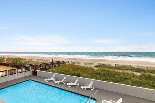 A rectangular outdoor pool with white lounge chairs overlooks a sandy beach and ocean under a clear blue sky.