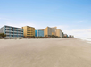 A row of beachfront hotels and buildings stands along a sandy, empty beach under a clear blue sky.