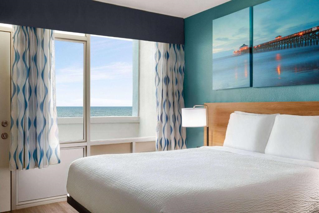 A neatly made bed in a modern hotel room with teal walls, large window showing an ocean view, patterned curtains, and wall art of a pier over the bed.