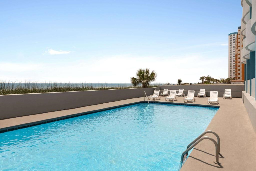 Outdoor swimming pool with clear blue water, surrounded by white lounge chairs, overlooking the ocean and a distant high-rise building under a clear sky.