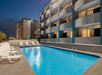Outdoor swimming pool beside a modern hotel building at dusk, with several white lounge chairs lined up along the pool deck.