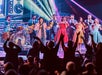 A band performs on stage with singers and musicians while an enthusiastic audience raises their hands. Bright stage lights and colorful outfits are visible.