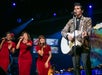 A man in a leopard print jacket plays guitar and sings on stage beside three women in red dresses, each holding a microphone. Stage lights are visible in the background.