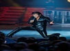 Performer in a black leather outfit kneels on stage while singing into a microphone under stage lights, with an audience watching.