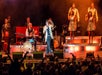 A band performs on stage with a singer at the front, backup singers, instrumentalists, and an enthusiastic audience in front of them.