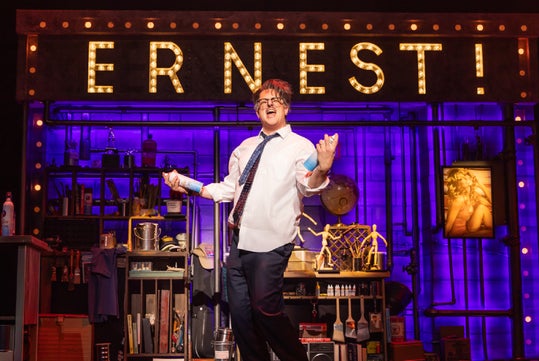 A performer in a white shirt and tie stands singing on a stage set with a lit "ERNEST!" sign and various props in the background.