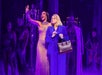 Two women stand center stage; one in a sparkling gown gestures dramatically, while the other in a blue suit holds a handbag. Several performers in dark outfits stand in the background amid purple lighting.