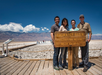 Four people stand smiling behind a sign that reads "Badwater Basin, 282 Feet/855 Meters Below Sea Level" on a sunny day, with a mountainous landscape in the background.