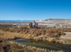 Four people stand near a small water stream amidst a desert landscape with mountains in the background under a clear blue sky.