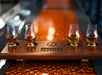 Four whiskey glasses with amber liquid are arranged on a wooden tasting flight board labeled "Decision Tactical," on a textured orange surface.