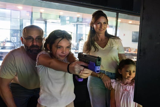 A young girl aims a purple training gun at a target range, supervised by an adult man, with a woman and another young girl standing beside them.