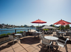 Rooftop patio with empty tables and chairs under red umbrellas, overlooking a body of water and city skyline on a clear, sunny day.