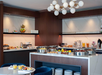 A modern breakfast buffet with various plated foods, chafing dishes, drinks, and fruit on a marble counter in a well-lit dining area with wooden and marble decor.