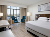Hotel room with a large bed, two blue chairs, and a desk. The room has a window with a view of water and city buildings. Neutral tones and modern decor.