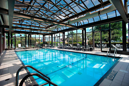 Indoor swimming pool with a glass roof and walls, surrounded by lounge chairs and tables. Greenery is visible outside the windows.