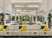 Spacious hotel lobby with white walls, circular ceiling lights, and gray seating. Cushions in blue and yellow accent the decor. Large windows and indoor plants provide a bright, airy feel.