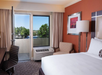 Hotel room with a bed, desk, chair, and artwork on the wall. A window and door open to a balcony with a view of trees and blue sky.