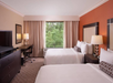 Hotel room with two double beds, a desk with a chair, a TV on a dresser, lamps, and a large window with a view of greenery.
