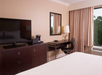 Hotel room with a bed, dark wood dresser, flat-screen TV, desk with a chair, a lamp, and a coffee maker. Window with closed curtains on the right.