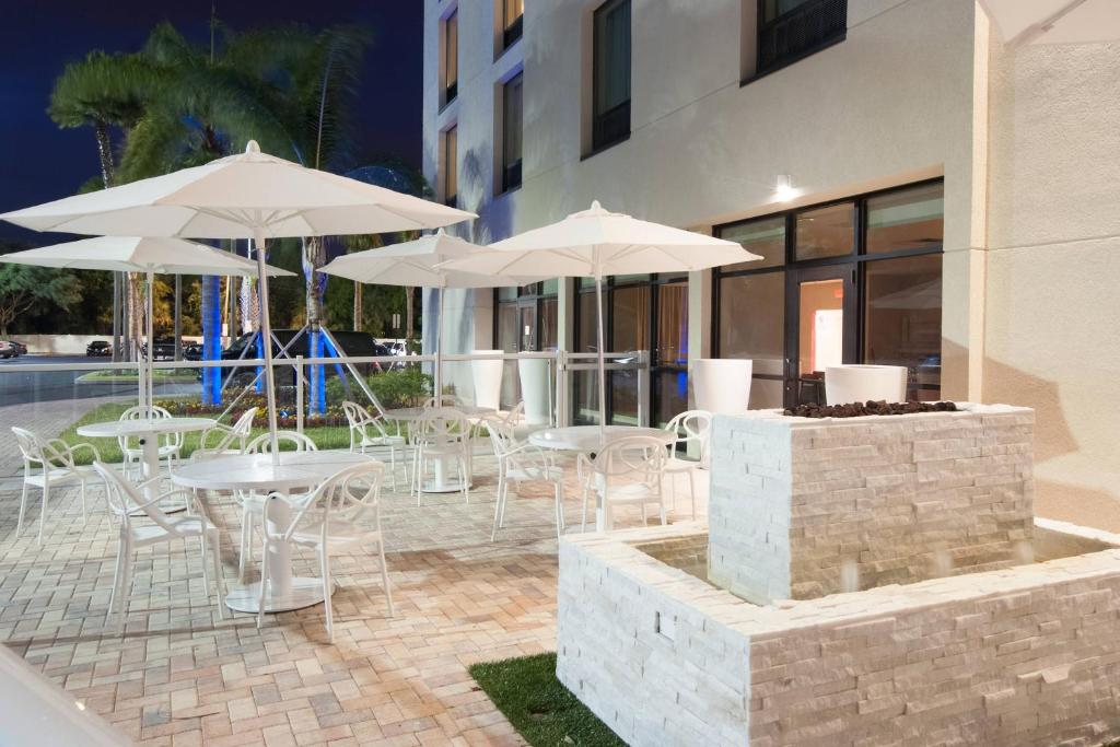 Outdoor seating area at night with white tables, chairs, and umbrellas on a tiled patio. A modern stone water fountain is in the foreground. Palm trees and a building are in the background.