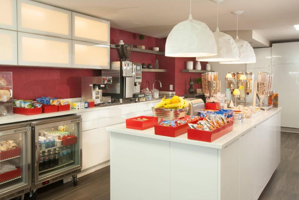 Hotel breakfast bar with an assortment of pastries, snacks, and a fruit basket. Beverage dispensers and a refrigerator with drinks are also visible. White cabinets and red accents decorate the area.