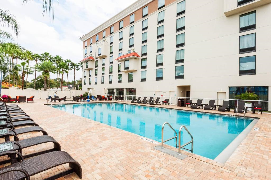 Outdoor hotel pool with lounge chairs on a brick patio, surrounded by a tall building and palm trees.