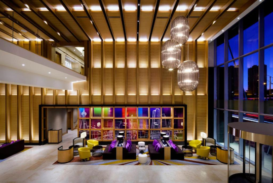 Modern hotel lobby with wooden walls, colorful seating areas, and decorative lighting fixtures. Large windows offer an expansive city view.