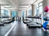 A gym with treadmills, exercise bikes, and colorful stability balls. Large windows and mirrors line the walls, providing ample natural light.