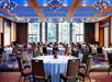 Ballroom with round tables and chairs set up for a formal event, featuring blue ceiling lights and large windows with an urban view.