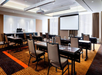 Conference room with rows of tables and chairs, a projector screen, and a refreshment station in the back. Natural light filters through full-length curtains.